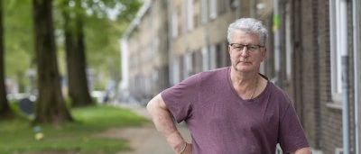 Man met grijs haar in een paarse shirt leunt tegen een hek in een stedelijke omgeving.