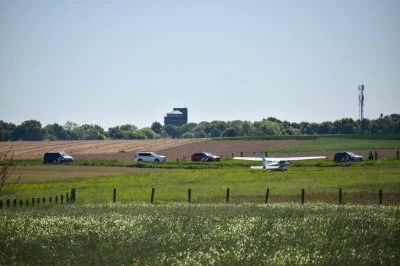 Een sportvliegtuigje staat stil aan het einde van een landweg met auto's van de hulpdiensten ernaast en groene velden op de achtergrond.