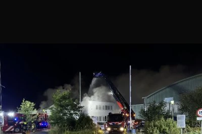 Brand op een industrieel terrein, met brandweermensen en rookpluimen in de lucht.