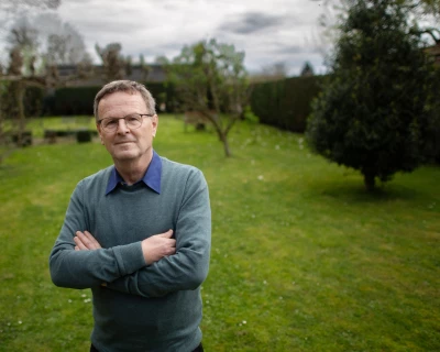 Man met kruisende armen in een groene tuin onder een bewolkte lucht.