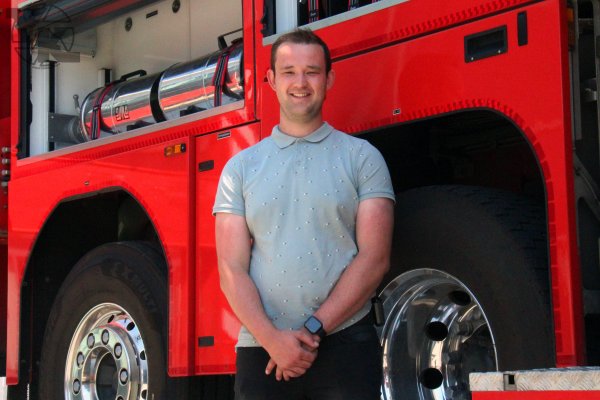 Jonge man in een lichtblauw shirt staat naast een rode brandweerwagen.